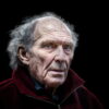 Elderly man with gray hair and a weathered face, wearing a dark red jacket, staring ahead with a serious expression, set against a black background. Elderly man with gray hair and a weathered face, wearing a dark red jacket, staring ahead with a serious expression, set against a black background.