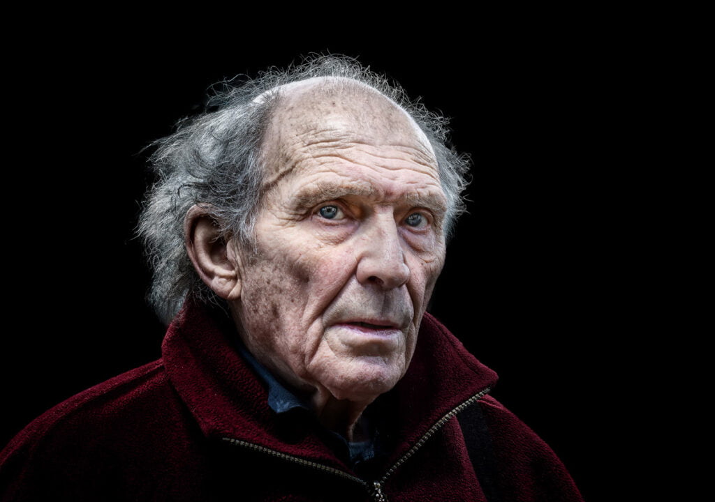 Elderly man with gray hair and a weathered face, wearing a dark red jacket, staring ahead with a serious expression, set against a black background.