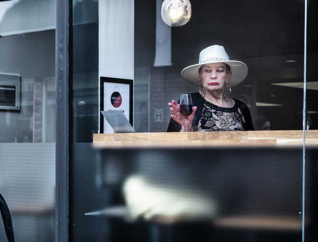 A woman wearing a white hat and black dress holds a glass of red wine, looking pensive, while standing inside a building by a window.