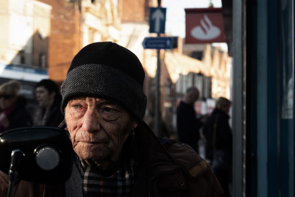 Elderly man wearing a beanie and scarf stands outdoors on a sunny day, with a group of people and buildings in the background.