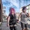 Two people with colorful punk hairstyles and clothing walk hand-in-hand on a city street during the day. They are dressed in leather and casual attire. Two people with colorful punk hairstyles and clothing walk hand-in-hand on a city street during the day. They are dressed in leather and casual attire.