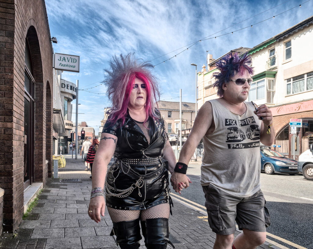 Two people with colorful punk hairstyles and clothing walk hand-in-hand on a city street during the day. They are dressed in leather and casual attire.