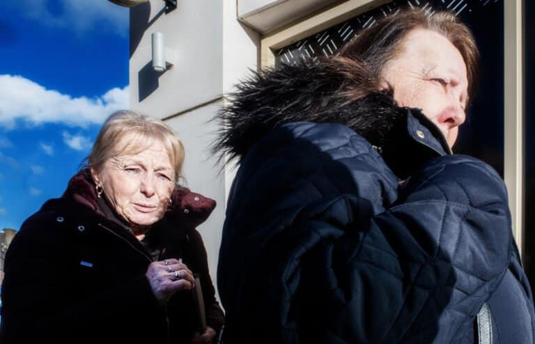Two women in dark winter jackets walk outside on a sunny day with a blue sky and partly cloudy background. The woman in front holds her collar, while the woman behind follows closely.