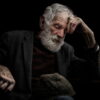 Elderly man with a white beard and mustache, wearing a dark jacket, resting his head on his hand while sitting and looking down, against a dark background. Elderly man with a white beard and mustache, wearing a dark jacket, resting his head on his hand while sitting and looking down, against a dark background.