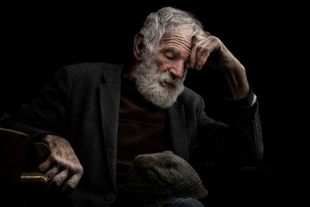 Elderly man with a white beard and mustache, wearing a dark jacket, resting his head on his hand while sitting and looking down, against a dark background.