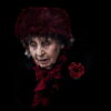 An elderly woman wearing a dark red fur hat, a black fur coat, and a red scarf, looks downward with a solemn expression against a black background. An elderly woman wearing a dark red fur hat, a black fur coat, and a red scarf, looks downward with a solemn expression against a black background.