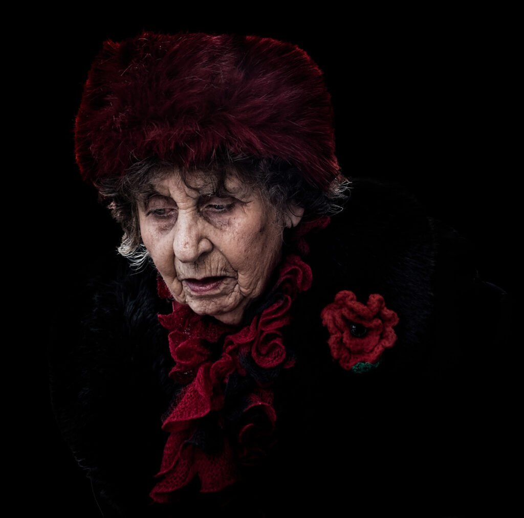 An elderly woman wearing a dark red fur hat, a black fur coat, and a red scarf, looks downward with a solemn expression against a black background.