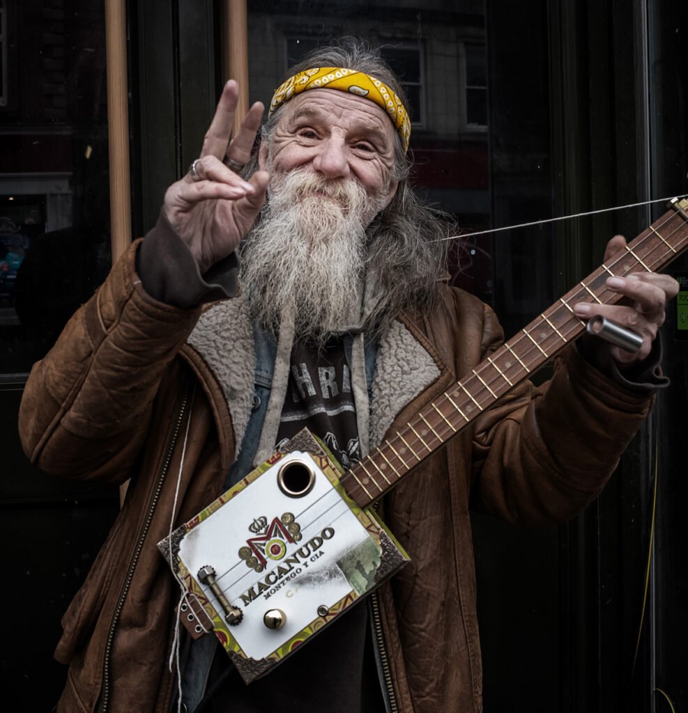Older man with a long beard and wearing a headband smiles and flashes a peace sign while holding a homemade guitar made from a cigar box. He is dressed in layered clothing, including a brown jacket.