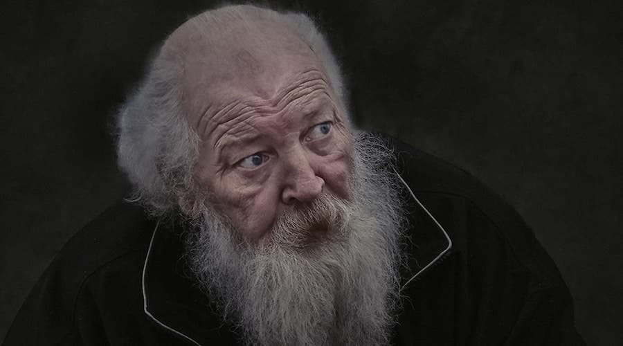 Elderly man with a long white beard and thinning hair, wearing a dark jacket, looking slightly upward with a serious expression against a dark background.
