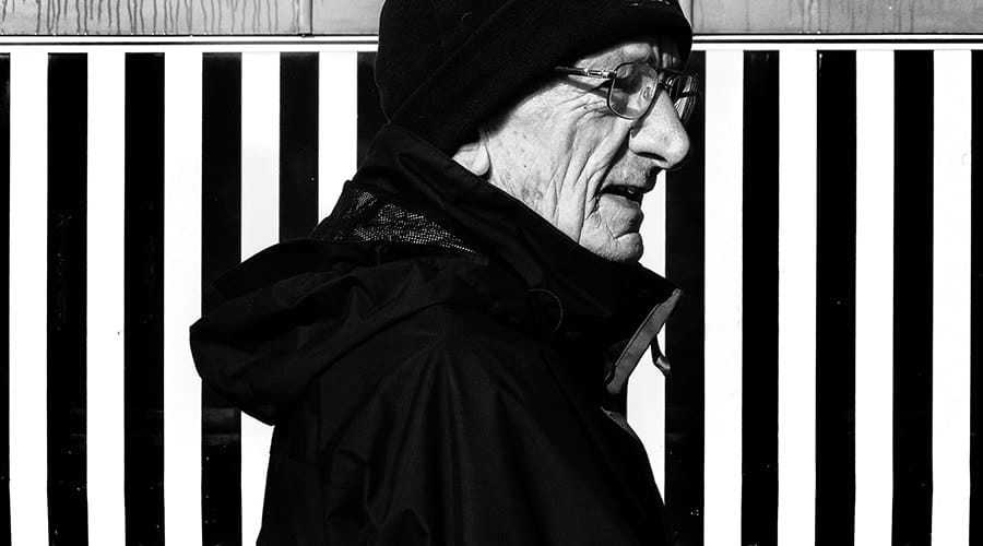 Older man in profile wearing glasses, a beanie, and a jacket, standing in front of a background with vertical black and white stripes.