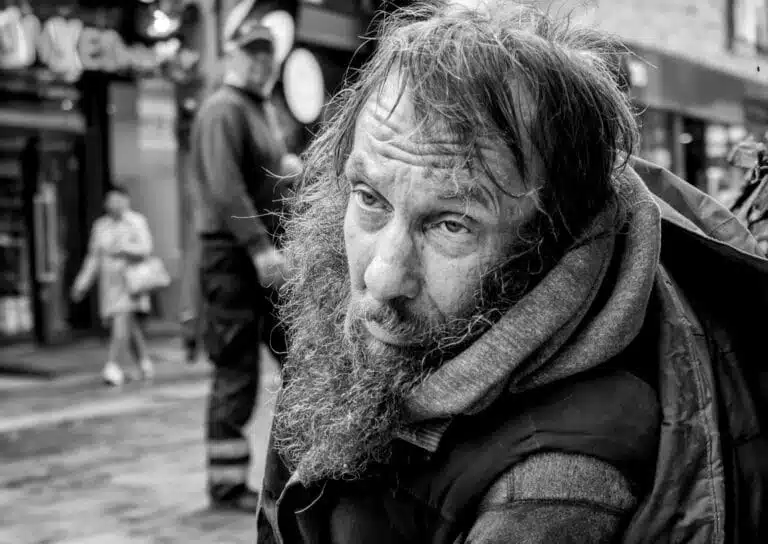 A black and white photo of a bearded man wearing a hood in an urban setting, with people walking in the background. A black and white photo of a bearded man wearing a hood in an urban setting, with people walking in the background.
