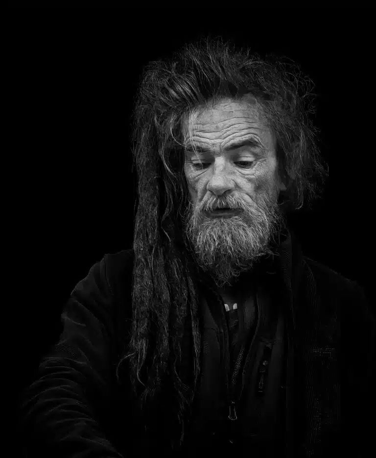 Black and white portrait of an older man with long, dreadlocked hair and a beard, wearing a dark jacket, against a black background. Black and white portrait of an older man with long, dreadlocked hair and a beard, wearing a dark jacket, against a black background.