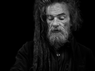 Black and white portrait of an older man with long, dreadlocked hair and a beard, wearing a dark jacket, against a black background.