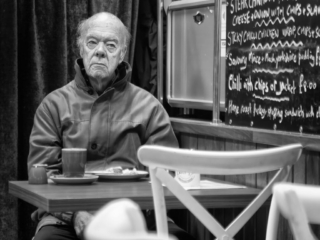 An older man sits alone at a caf&eacute; table with a mug and plate in front of him. In the background, a menu is displayed on a chalkboard. The scene is in black and white.