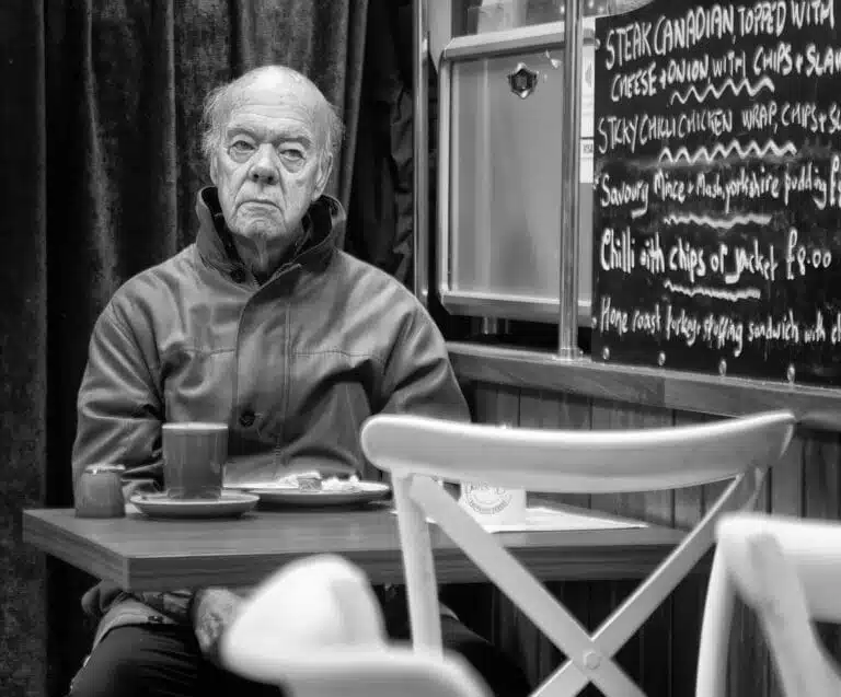 An older man sits alone at a café table with a mug and plate in front of him. In the background, a menu is displayed on a chalkboard. The scene is in black and white. An older man sits alone at a café table with a mug and plate in front of him. In the background, a menu is displayed on a chalkboard. The scene is in black and white.