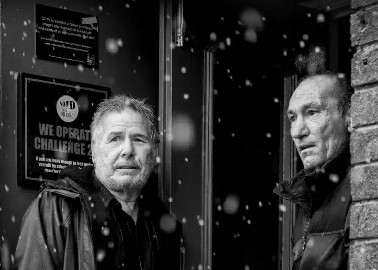 Two men stand at a doorway, looking out into falling snow. They are wearing jackets, and signs are visible on the wall beside them. The image is in black and white. Two men stand at a doorway, looking out into falling snow. They are wearing jackets, and signs are visible on the wall beside them. The image is in black and white.