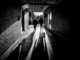 Two people walk through a dimly lit brick tunnel, casting long shadows on the ground.