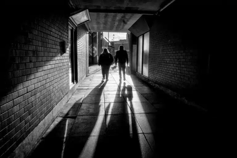 Two people walk through a dimly lit brick tunnel, casting long shadows on the ground. Two people walk through a dimly lit brick tunnel, casting long shadows on the ground.
