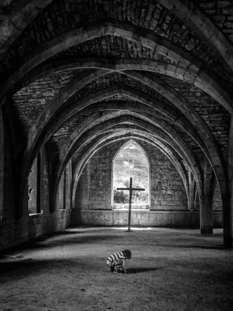 A child crouches under gothic stone arches in a dimly lit room, with a large window and cross in the background. A child crouches under gothic stone arches in a dimly lit room, with a large window and cross in the background.