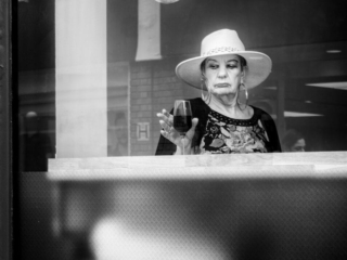 Woman in a hat stands behind a glass window, holding a glass, with a light bulb above her. Black and white image.
