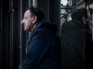 A man with a weathered face and disheveled hair stands next to a building, looking to the side. His reflection is visible in the dark glass beside him.