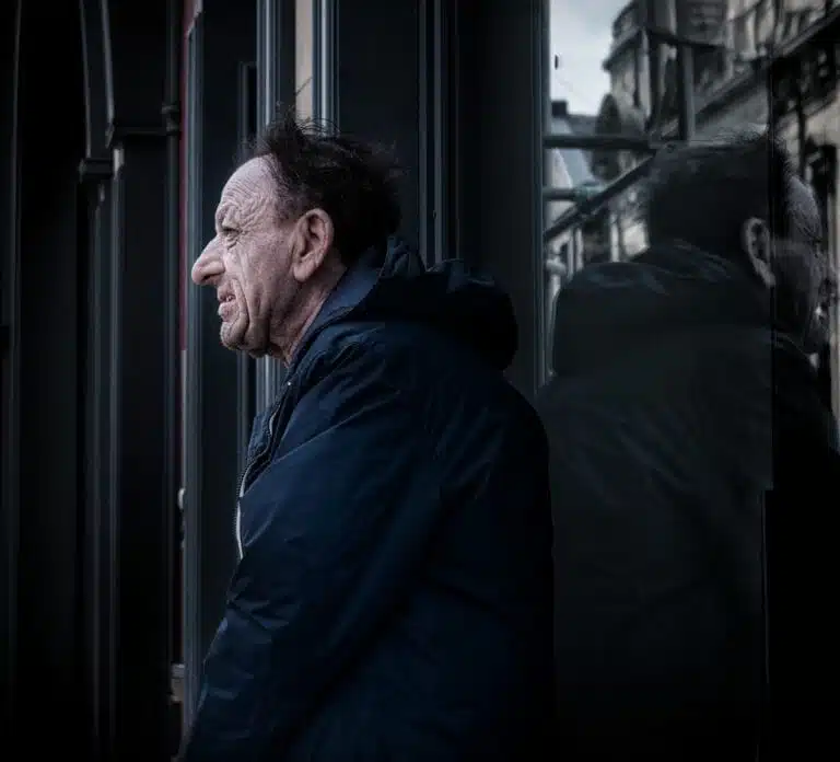 A man with a weathered face and disheveled hair stands next to a building, looking to the side. His reflection is visible in the dark glass beside him. A man with a weathered face and disheveled hair stands next to a building, looking to the side. His reflection is visible in the dark glass beside him.