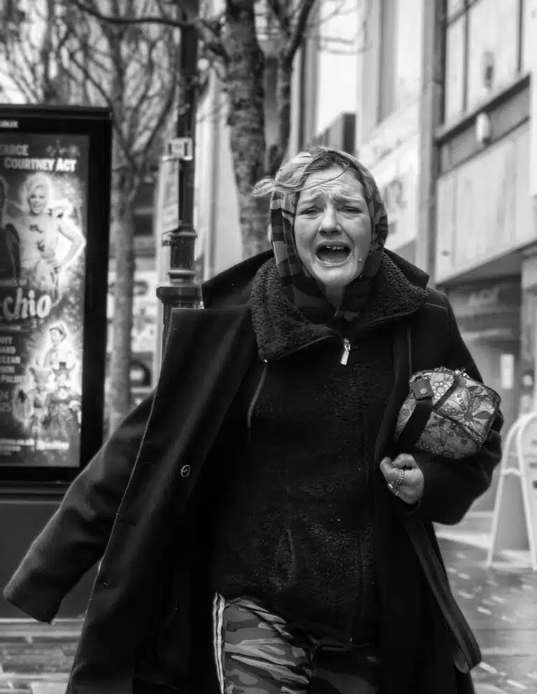A person wearing a coat and scarf is walking while holding a small bag. They appear to be talking or shouting. Street signs and a poster are visible in the background. A person wearing a coat and scarf is walking while holding a small bag. They appear to be talking or shouting. Street signs and a poster are visible in the background.