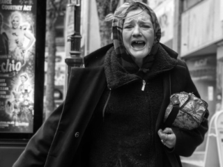 A person wearing a coat and scarf is walking while holding a small bag. They appear to be talking or shouting. Street signs and a poster are visible in the background.