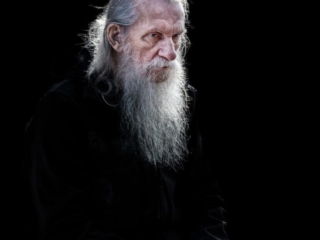 A man with long white hair and a beard sits against a black background, holding an object.