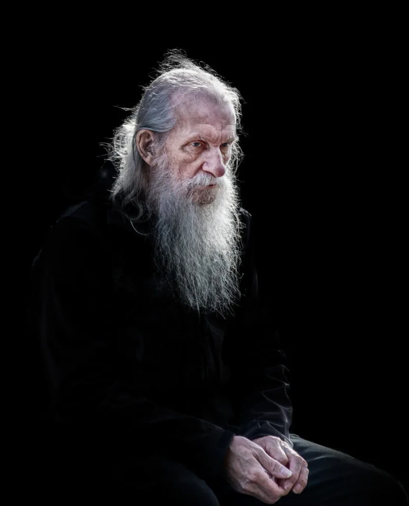 A man with long white hair and a beard sits against a black background, holding an object.