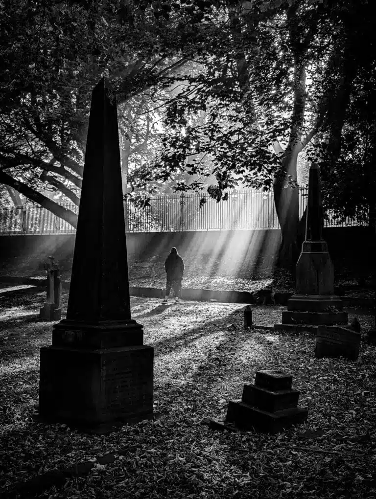 A silhouette of a person stands near gravestones in a cemetery, with sunlight filtering through the trees. A silhouette of a person stands near gravestones in a cemetery, with sunlight filtering through the trees.