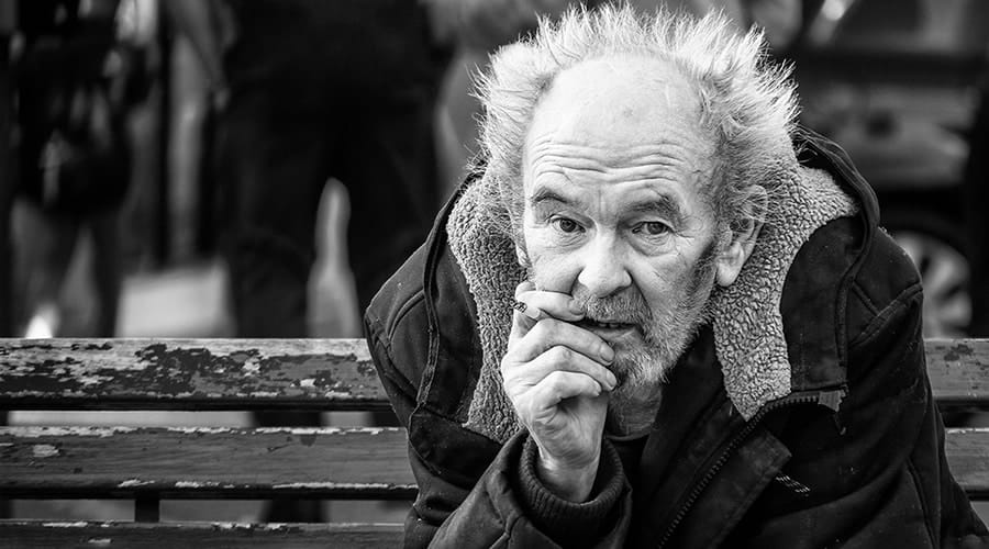 An older man with messy hair sits on a worn bench, wearing a heavy jacket, resting his hand near his face, looking directly at the camera. The photo is black and white.