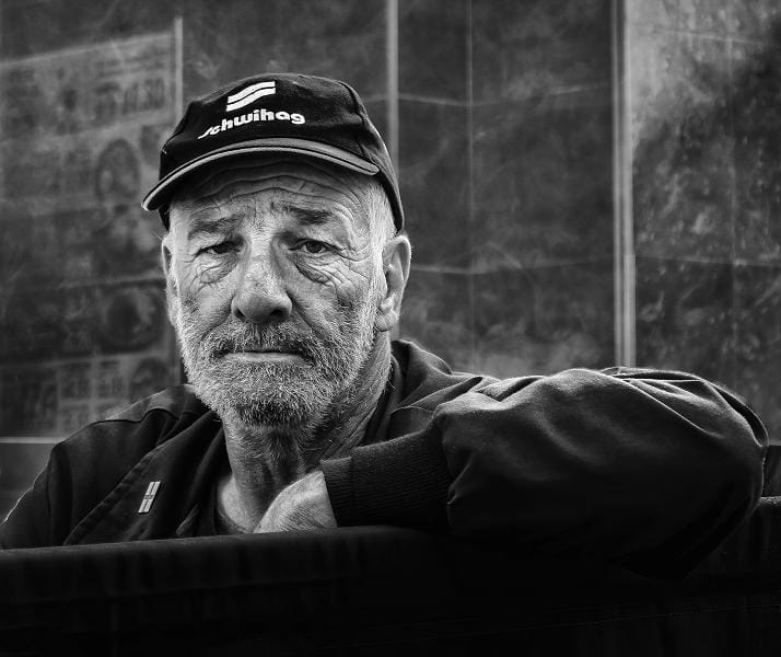 Elderly man with a weathered face and beard, wearing a cap and jacket, rests his arm on a dark surface; background is blurred cityscape.