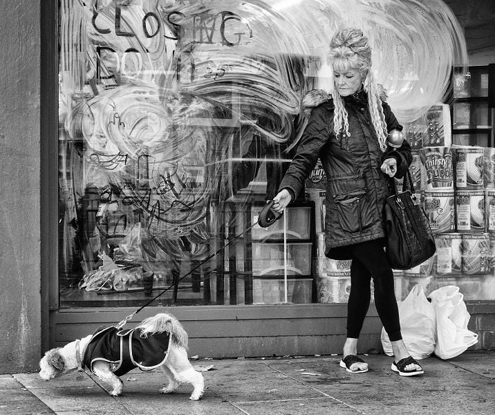A woman with a beehive hairstyle walks a small dog on a leash past a shop window with "CLOSING DOWN" painted on it. Plastic bags and paint cans are visible nearby.