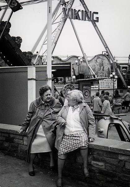 Two older women sit on a low wall having a conversation at an amusement park, with a Viking ship ride and other attractions visible in the background.