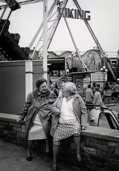 Two older women sit on a low wall having a conversation at an amusement park, with a Viking ship ride and other attractions visible in the background.