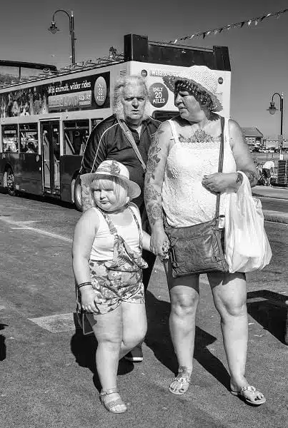 Three people, two adults and one child, stand on a sidewalk near a bus on a sunny day. The adults wear summer clothes; the child wears a hat and patterned romper.