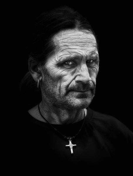 Middle-aged man with long hair, facial hair, and visible wrinkles, wearing an earring, black shirt, and a necklace with a cross, set against a dark background.