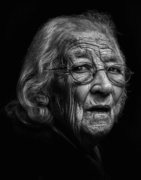 Black and white portrait of an elderly person with long gray hair and glasses, looking over their shoulder with a neutral expression. Wrinkles and facial details are prominently visible.